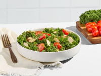 A bowl of salad on a white background next to a cutting board with kale and tomatoes.