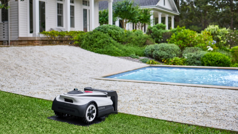 A robotic lawn mower resting on a lush green lawn near a pool, surrounded by a well-manicured garden and a white modern house in the background.
