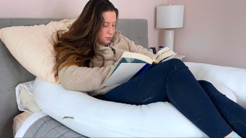 A woman sitting in bed reading a book, using the Hugl body pillow for support.