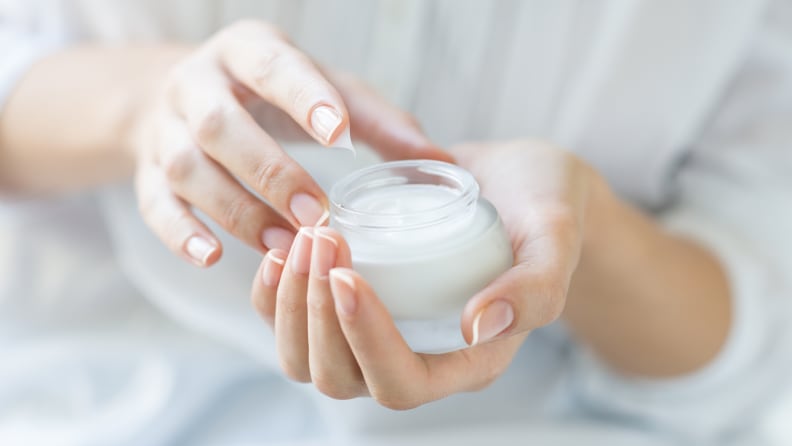 A person holds a jar of moisturizer in one hand and dips their other hand into the jar.