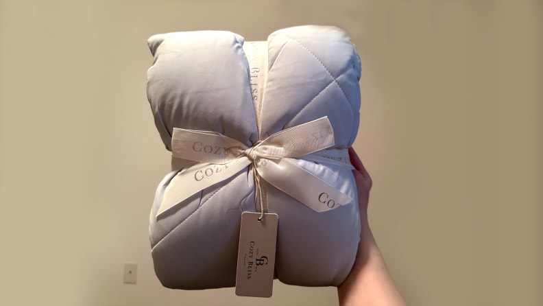 Gray Cozy Bliss comforter being held in front of a beige wall, folded into a square cushion shape tied together with a white ribbon.