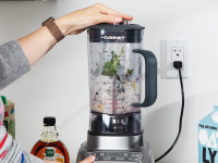 The Cuisinart blender sits on a counter and a person has their hand on the lid and a finger on the control panel.