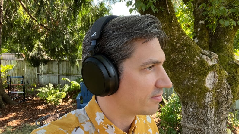 A man in a yellow pattern shirt listens to a black pair of headphones in a green yard.