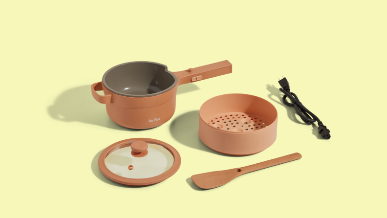 The components of the Perfect Power Pot on a yellow background, including the pot base, lid, steamer attachment, spatula, and cord.