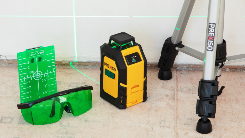 A yellow laser next to goggles and a tripod shoots a green laser onto a wall.