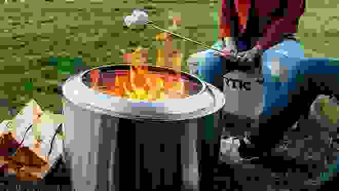 A person roasting a marshmellow over a Solo stove in a backyard grass environment.