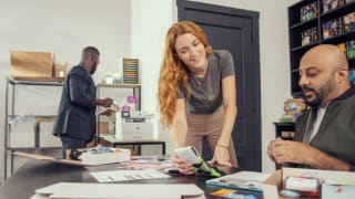 Woman holding patterned socks up for a coworker while another man uses the printer in the background