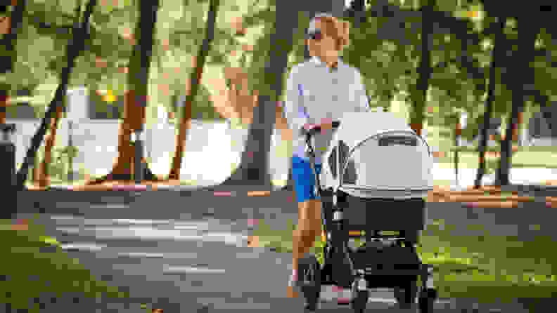 A woman in a white shirt and sunglasses pushes a covered strolled through a park on a sunny day.