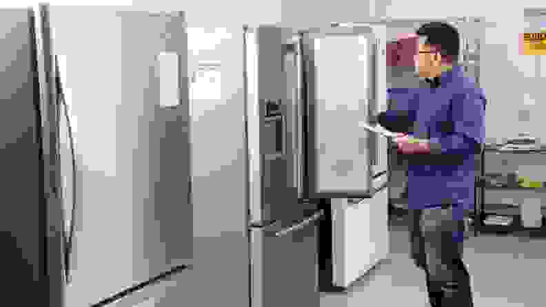 A man stands in a fridge testing lab with a clipboard in hand, in front of a row of refrigerators. He's holding one of their doors open, examining the interior.