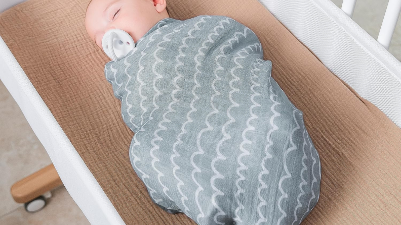 Overhead view of a baby sublimely sleeping while wrapped in a gray Comfy Cubs swaddle blanket in a crib.