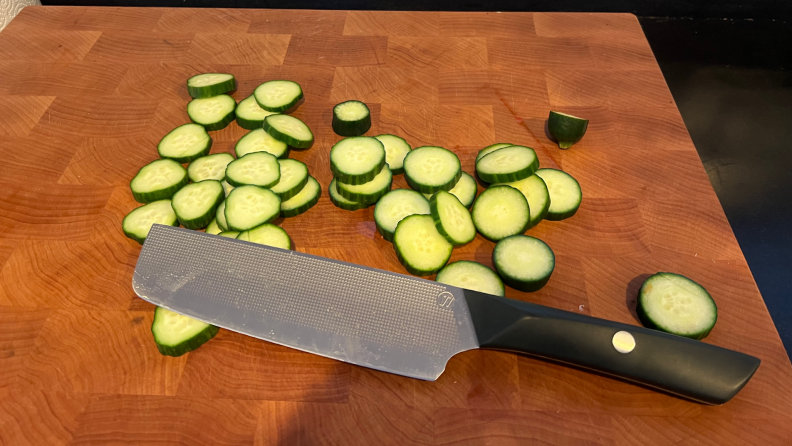 A Nakiri knife sitting next to a pile of cucumber slices sitting on top of a wooden cutting board.