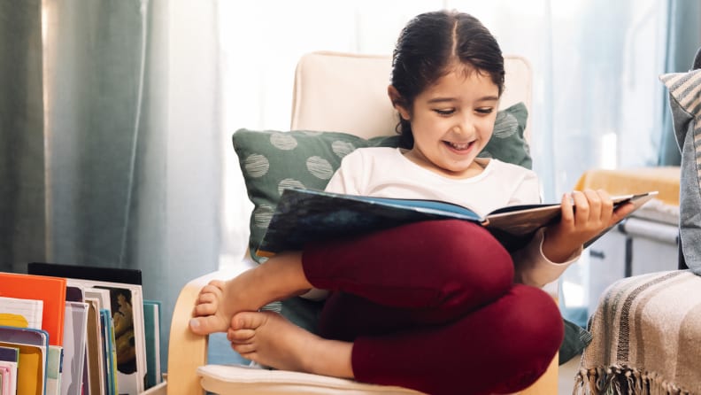 Young girl smiling while reading a large picture book in chair next to books with legs and feet tucked next to her.