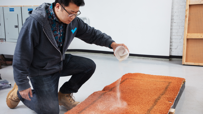 A man pouring a white powder on an orange carpet to test vacuums with