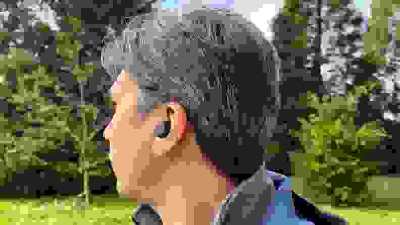 An earbud sits in the ear of a man with brown and white hair in a park with trees and grass.