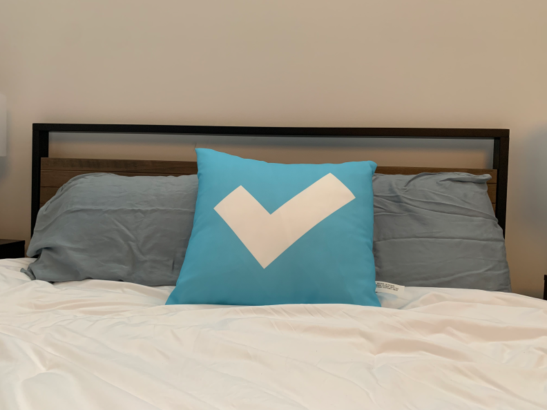 Three blue pillows on a white bed. The first pillow is bright blue with a white check mark. The two pillows behind it are lighter blue.