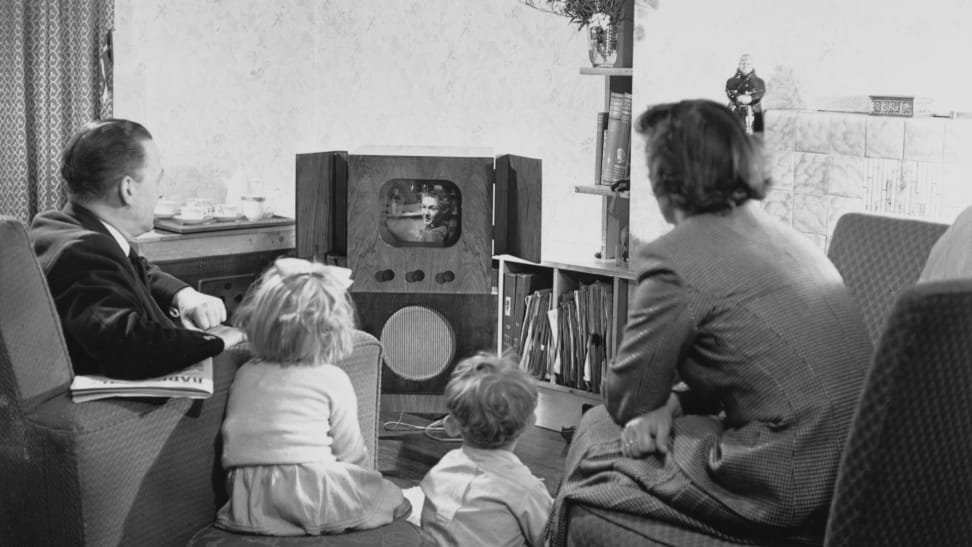 A family of four - a mother, father, and two small children - sit around a small black-and-white television screen in the 1950s.