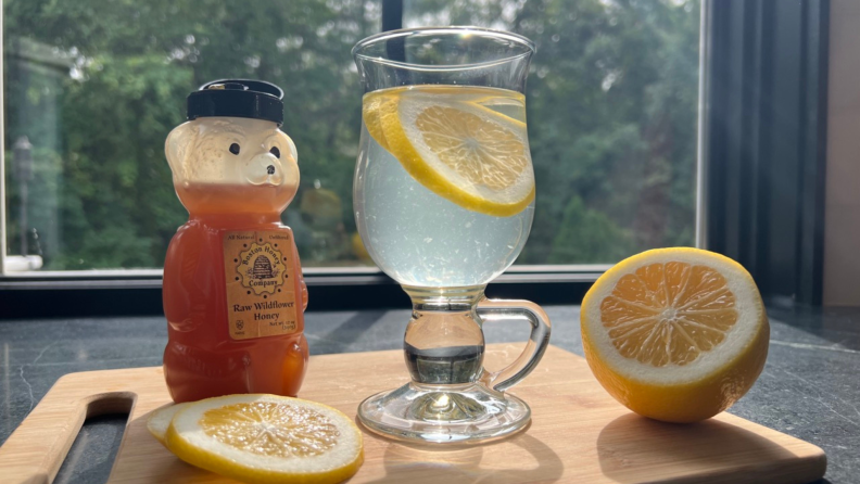 A bear bottle of honey sits behind lemon slices next to a glass mug of water with lemon, and a half a lemon on a cutting board.