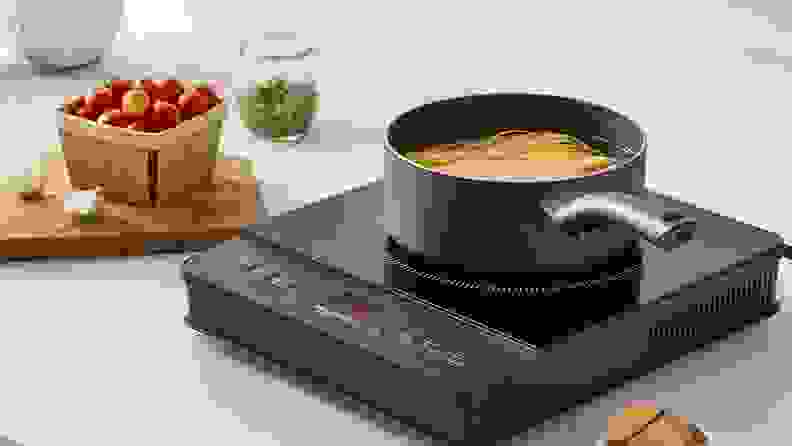 A black hot plate sits on a kitchen countertop as a pot of pasta and boiling water cooks on top of it.
