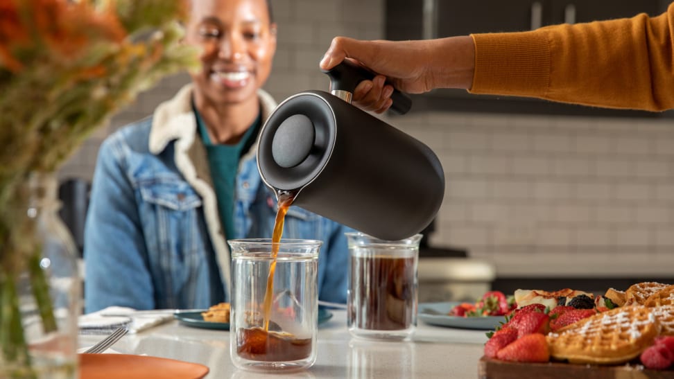 A person is pouring coffee from a Fellow Clara French press coffee maker into a glass mug. A different person is smiling at the mug, watching the coffee pour.