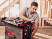 A model in a gray T-shirt demonstrates a Skil table saw at a construction site.