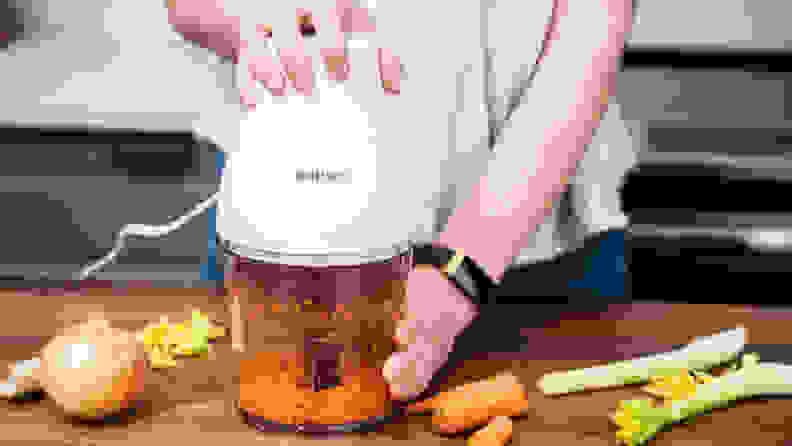 A person is pressing down a mini food processor to get it to work, which seems to be chopping carrots.