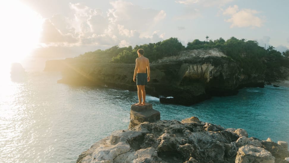 Man standing on a cliff in front of the ocean wearing blue swim trunks