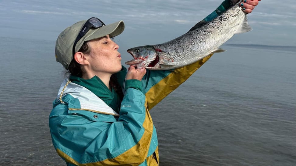 A woman wearing an Eddie Bauer jacket is holding a fish and pretending to kiss the fish.