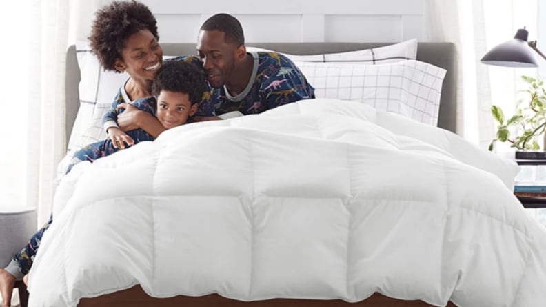 A Black mom, dad, and child hugging in a bed covered by a white down comforter.