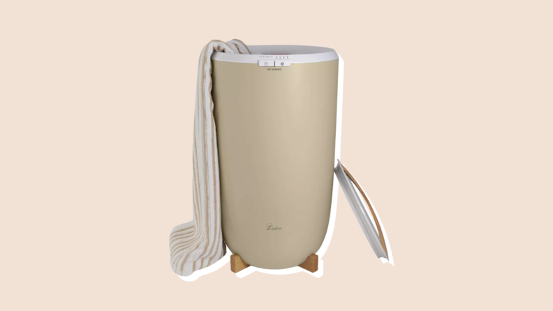 A tan bucket with white top ring and controls heats a long bath towel while sitting on a wooden base with the lid leaning on it.