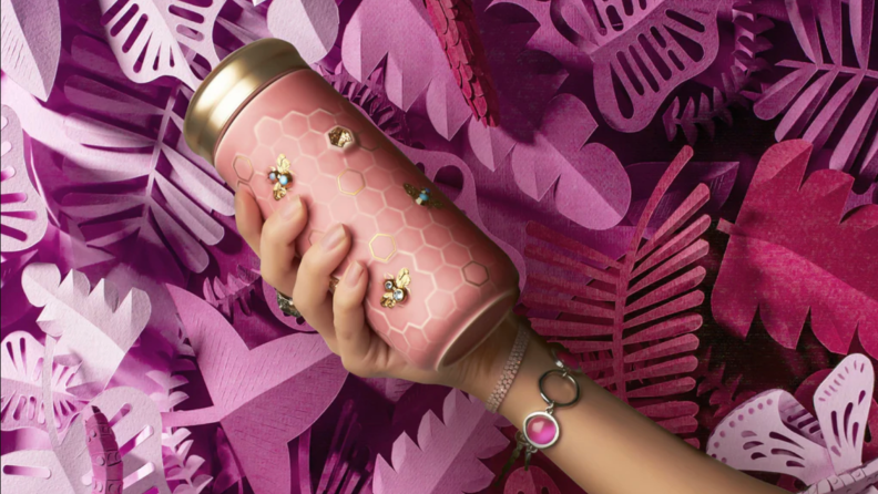 A tan hand holds a peach water bottle on a purple frilly background