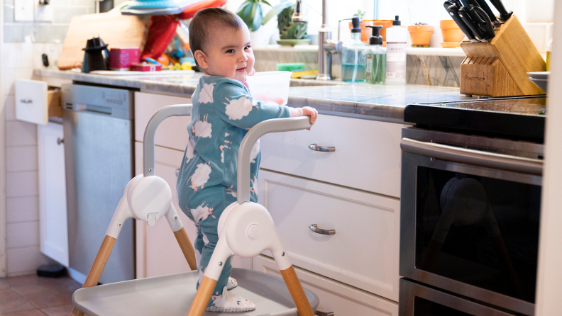 A toddler standing on the base of the Skip Hop Sit-to-Step high chair playing in the kitchen sink.