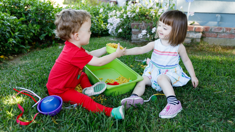 Young boy and girl playing outdoors with a sensory bin filled with uncooked pasta.