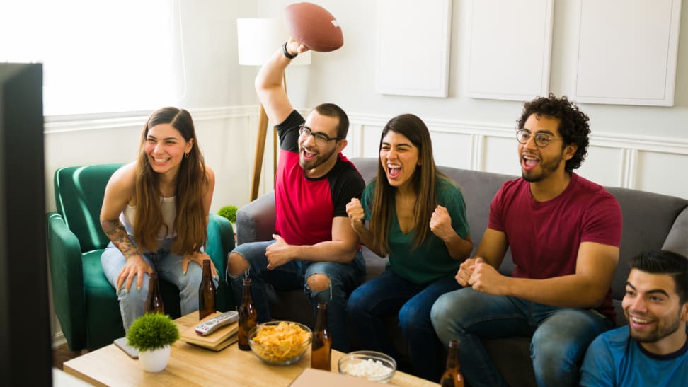 Five people sitting on a couch in the living room watching football on a flat screen TV