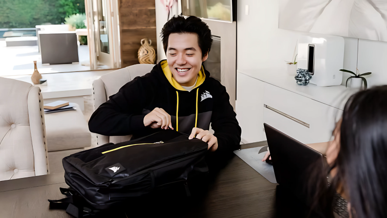A young man smiles while unzipping a Corsair backpack at a table during a casual work-from-home setup.