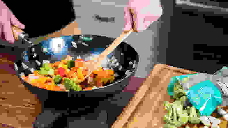 A person is using a spatula to stir a medley of vegetables in a carbon steel wok.