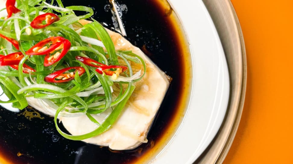 A white plate with steamed white fish topped with soy sauce, green onions, whole ginger, and limes.