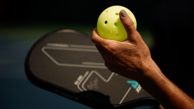 Close-up of a left hand on a yellow-green pickleball, a black paddle in the right hand, as a player prepares to serve.