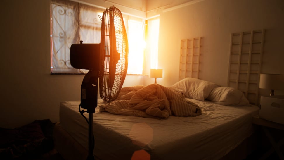 A bedroom with a messy bed, the sun shining through the window, and the sillhouette of a fan.