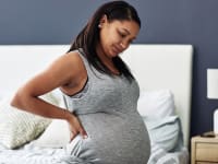 Pregnant person sitting on bed with hand on back in discomfort.