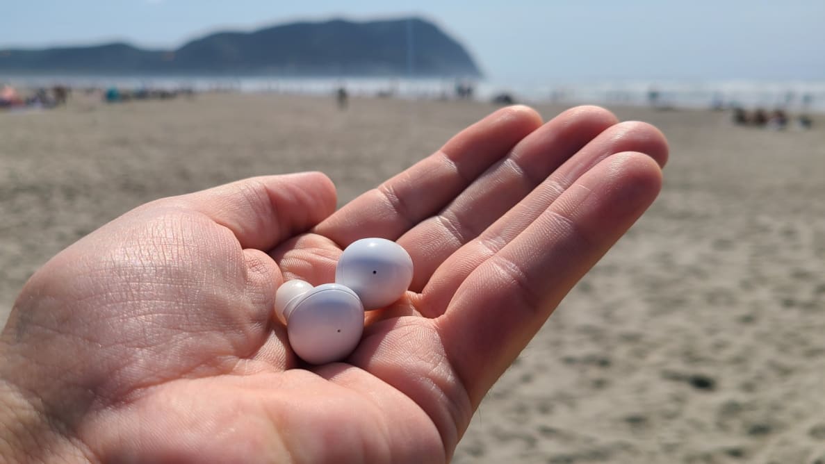 The all white Galaxy Buds 2 sit together in a hand raised with a sandy beach and mountains in the background.