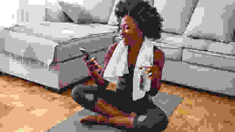 Woman using smart phone on exercise mat in front of her laptop. Sporty girl is exercising at phone on mat enjoying playlist on mobile phone. Positive active energtic sportswoman sit on purple mat use cellphone
