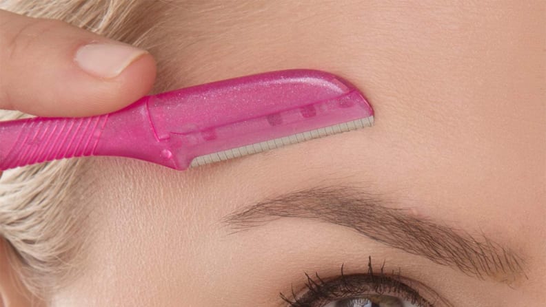 A closeup of a woman holding a dermaplane tool above her eyebrow pointing down.