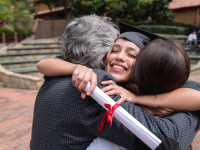 Girl smiling wearing cap and gown hugs parents while holding a diploma in hand.