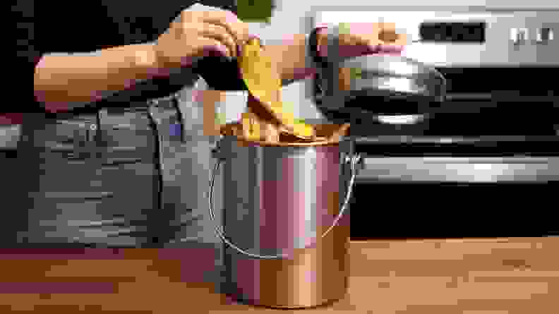 Person placing brown banana peel in silver can on wooden countertop surface in kitchen.