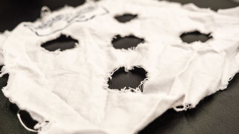 A mechanical action strip–a small square swatch of linen with five holes punched in it like face of a die—lies on a surface. The threads around the edges of the swatch and inside the punched holes is very frayed.