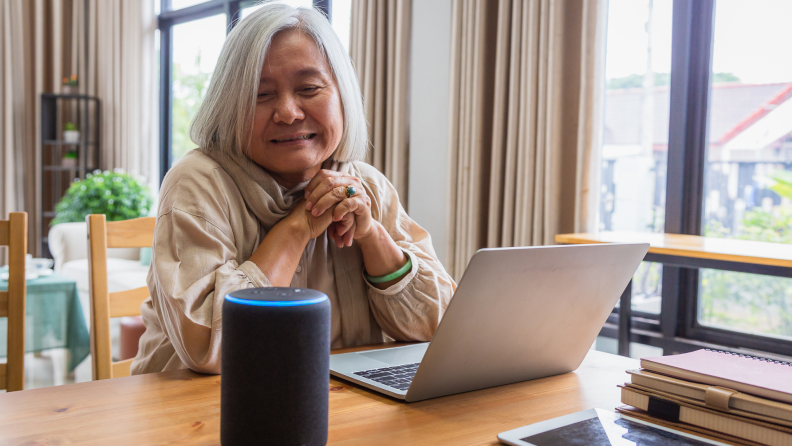 Senior woman sitting at table in front of laptop, next to an Alexa smart device at home.