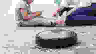 A Eufy 11S robot vacuum cleaning a carpet with a woman and child sitting in the background.