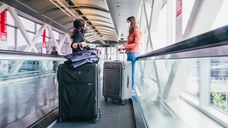 Two women stand on an airport people mover, holding on to their luggage