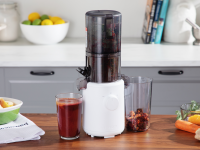 The Hurom H310A Slow Juicer sits on a wood counter in a kitchen with a full cup of juice by it, and bowls of produce on the counter.