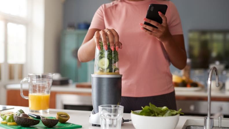 Eine Frau, die das Magic Bullet gedrückt hält, um einen Smoothie mit Produkten wie Grünkohl, Gurken und Kiwi zu mixen, während sie auf ihrem Telefon Multitasking betreibt.